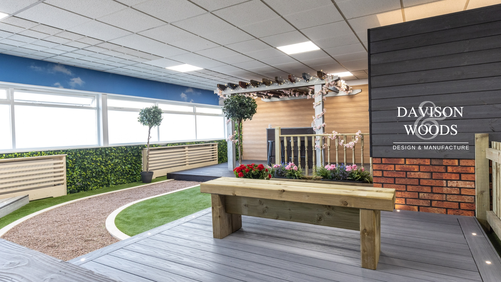 Grey timber decking and built in lights, in the centre, a light brown timber bench made from BSW railway sleepers. To the left, a pink gravel path leads onto the pergola section in the background.