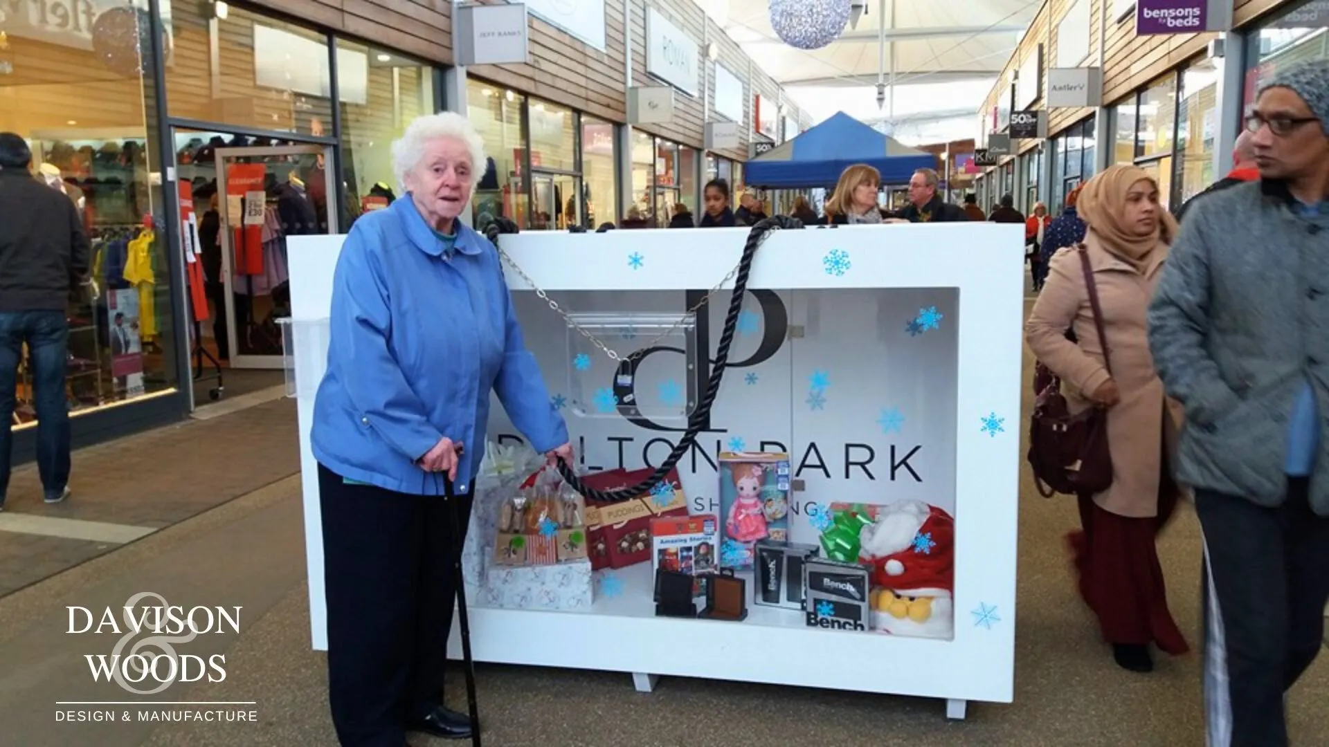 Woman standing next to the Display case in the shape of a white shopping back the front window shows presents inside.